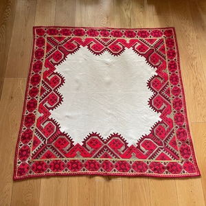 May include: A white tablecloth with a red and gold embroidered border. The border features a geometric pattern.