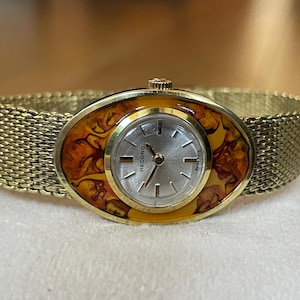 Op de afbeelding: Een ovaalvormig polshorloge in goudkleur met een witte wijzerplaat en zwarte wijzers. Het horloge heeft een goudkleurige mesh-band en een bezel met een bruin en oranje patroon. De wijzerplaat van het horloge heeft "Record" in zwarte letters.
