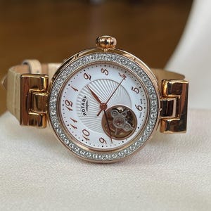 Op de afbeelding: Een roségouden Rotary-horloge met een witte wijzerplaat, zichtbaar uurwerk en een bezel bezet met kleine, heldere kristallen. Het heeft een lichtbeige leren bandje en wordt gepresenteerd op een wit oppervlak. De diameter van de wijzerplaat is ongeveer 3 cm.
