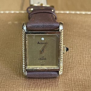 K&ouml;nnte beinhalten: Eine goldfarbene Accuriest-Uhr mit einem braunen Lederarmband. Das Zifferblatt der Uhr ist rechteckig und hat eine goldfarbene L&uuml;nette. Das Zifferblatt ist wei&szlig; mit schwarzen Zeigern und schwarzen r&ouml;mischen Ziffern als Stundenmarkierungen. Das Zifferblatt hat auch einen kleinen Diamanten an der 12-Uhr-Position. Auf dem Zifferblatt steht "Accuriest" und "17 Jewels".