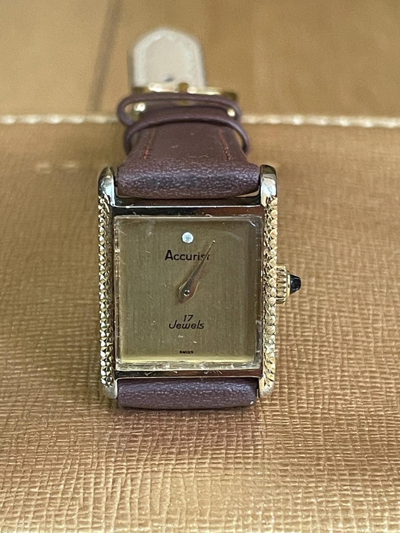K&ouml;nnte beinhalten: Eine goldfarbene Accuriest-Uhr mit einem braunen Lederarmband. Das Zifferblatt der Uhr ist rechteckig und hat eine goldfarbene L&uuml;nette. Das Zifferblatt ist wei&szlig; mit schwarzen Zeigern und schwarzen r&ouml;mischen Ziffern als Stundenmarkierungen. Das Zifferblatt hat auch einen kleinen Diamanten an der 12-Uhr-Position. Auf dem Zifferblatt steht "Accuriest" und "17 Jewels".