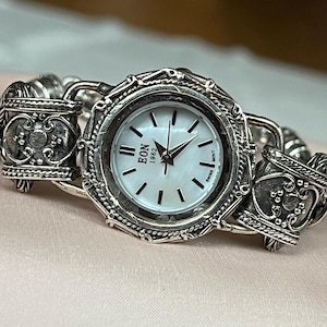 Könnte beinhalten: Eine silberfarbene Armbanduhr mit weißem Zifferblatt und schwarzen Zeigern. Die Uhr hat eine dekorative Lünette und ein Armband mit aufwendigen Details. Das Zifferblatt zeigt den Markennamen "BON 1962" und das Wort "SWISS".