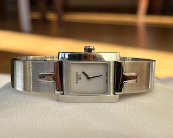 Peut inclure: Une montre Tissot argentée avec un cadran blanc rectangulaire et des aiguilles noires. Le bracelet est un large bracelet en métal brossé. Le cadran affiche le logo Tissot et le texte "SWISS MADE".