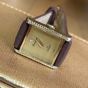 K&ouml;nnte beinhalten: Eine goldfarbene Accustar Armbanduhr mit einem braunen Lederarmband. Das Zifferblatt der Uhr ist quadratisch und hat eine rautenf&ouml;rmige Markierung an der 12-Uhr-Position. Das Zifferblatt der Uhr zeigt auch "Accustar" und "17 Jewels".