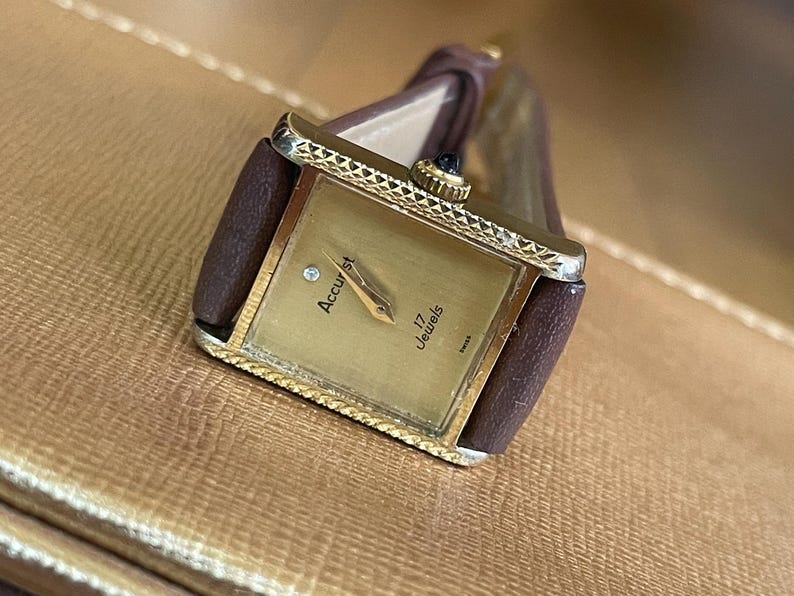 K&ouml;nnte beinhalten: Eine goldfarbene Accustar Armbanduhr mit einem braunen Lederarmband. Das Zifferblatt der Uhr ist quadratisch und hat eine rautenf&ouml;rmige Markierung an der 12-Uhr-Position. Das Zifferblatt der Uhr zeigt auch "Accustar" und "17 Jewels".