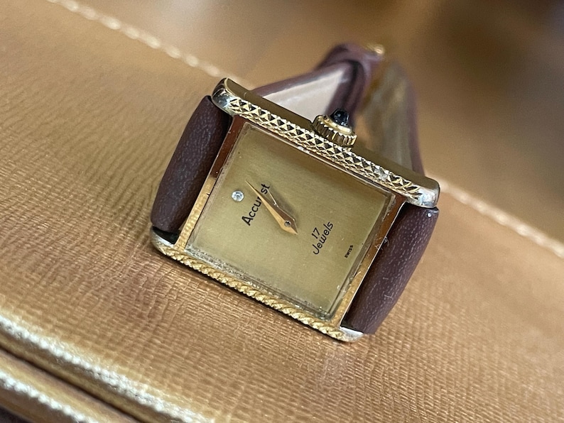 K&ouml;nnte beinhalten: Eine goldfarbene Accust-Uhr mit einem braunen Lederarmband. Das Zifferblatt der Uhr ist rechteckig und hat einen Diamanten an der 12-Uhr-Position. Das Zifferblatt der Uhr zeigt auch "Accust" und "17 Jewels".