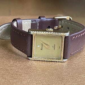 K&ouml;nnte beinhalten: Eine goldfarbene Accurif Armbanduhr mit quadratischem Zifferblatt und braunem Lederarmband. Das Zifferblatt hat einen Diamanten auf der 12-Uhr-Position und die Aufschrift "Accurif 17 Jewels".
