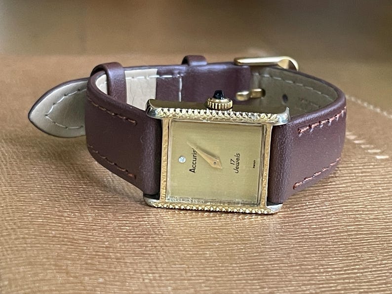 K&ouml;nnte beinhalten: Eine goldfarbene Accurif Armbanduhr mit quadratischem Zifferblatt und braunem Lederarmband. Das Zifferblatt hat einen Diamanten auf der 12-Uhr-Position und die Aufschrift "Accurif 17 Jewels".