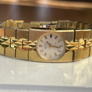 May include: A gold-toned wristwatch with a white face and black Roman numeral hour markers. The watch has a gold-toned metal band with a textured design. The watch face says "ROXY ANKER 17  RUBIS INCABLOC".