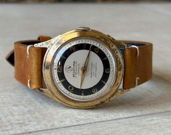 May include: Vintage Mortima Jeweled watch with a gold-tone case and a brown leather strap. The watch face is white and black with gold hands and numerals. The watch is marked "RESISTANT, DUST, ANTIMAGNETIC."