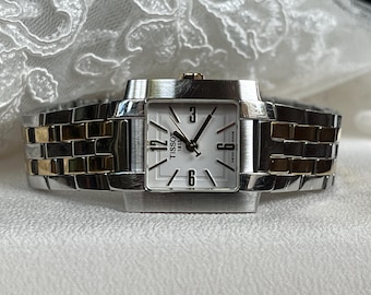 Peut inclure: Une montre Tissot argentée et dorée avec un cadran carré blanc. La montre a un bracelet argenté avec des accents dorés. Le cadran affiche les chiffres 12, 3, 6 et 9, avec le logo Tissot 1853.