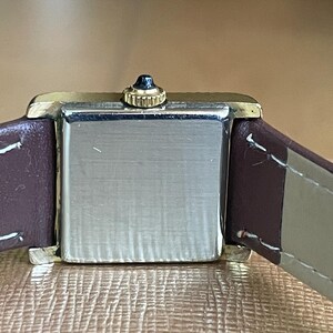 K&ouml;nnte beinhalten: Eine quadratische Armbanduhr mit einem braunen Lederarmband. Das Zifferblatt der Uhr ist silberfarben und hat eine goldfarbene L&uuml;nette.