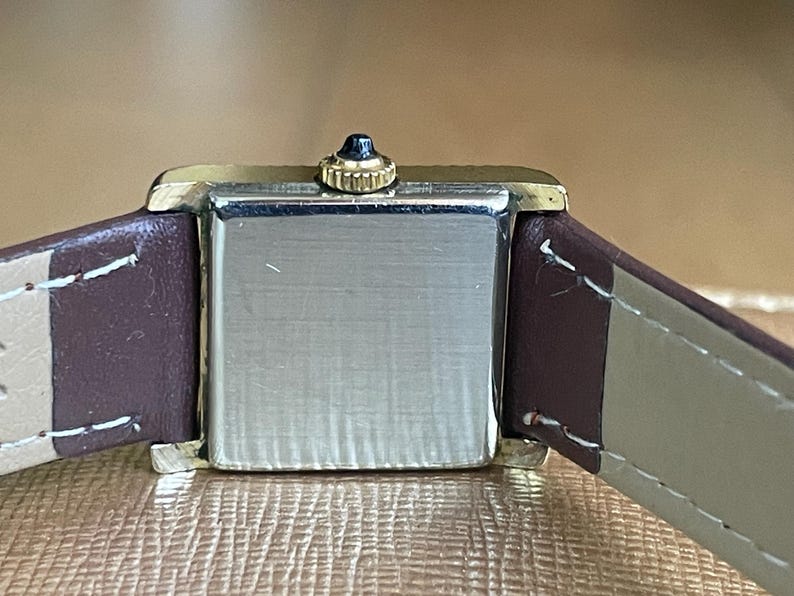 K&ouml;nnte beinhalten: Eine quadratische Armbanduhr mit einem braunen Lederarmband. Das Zifferblatt der Uhr ist silberfarben und hat eine goldfarbene L&uuml;nette.