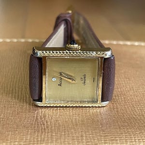K&ouml;nnte beinhalten: Eine goldfarbene Armbanduhr mit einem braunen Lederarmband. Das Zifferblatt der Uhr ist quadratisch und hat ein wei&szlig;es Zifferblatt mit schwarzen Markierungen. Der Text "Accurist 17 Jewels" ist auf dem Zifferblatt aufgedruckt.