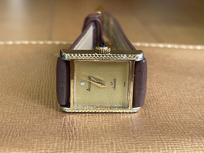 K&ouml;nnte beinhalten: Eine goldfarbene Armbanduhr mit einem braunen Lederarmband. Das Zifferblatt der Uhr ist quadratisch und hat ein wei&szlig;es Zifferblatt mit schwarzen Markierungen. Der Text "Accurist 17 Jewels" ist auf dem Zifferblatt aufgedruckt.