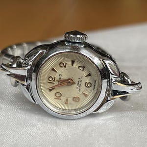 May include: A silver wristwatch with a white face and black numbers. The watch has a silver band and says "17 Jewels Antimagnetic" on the face.