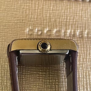 K&ouml;nnte beinhalten: Eine goldfarbene Armbanduhr mit einem braunen Lederarmband. Die Uhr hat ein quadratisches Zifferblatt und ein schwarzes Zifferblatt.