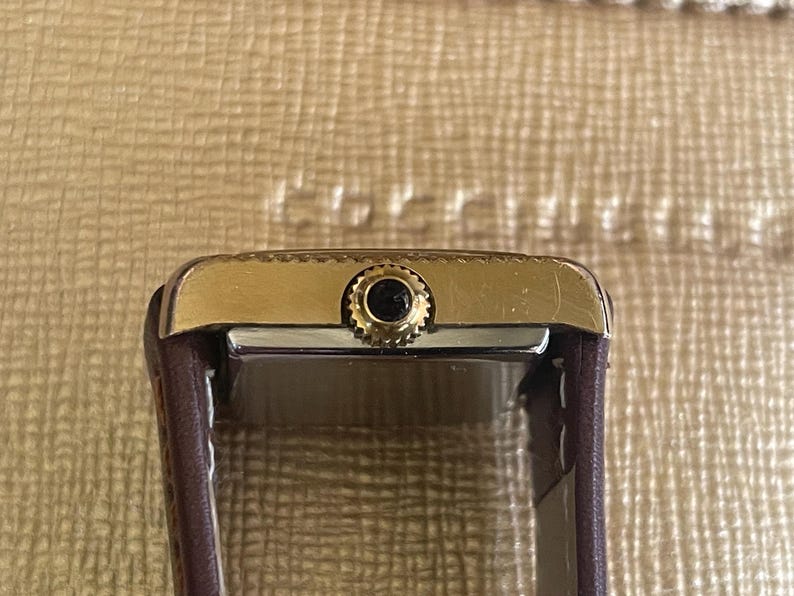 K&ouml;nnte beinhalten: Eine goldfarbene Armbanduhr mit einem braunen Lederarmband. Die Uhr hat ein quadratisches Zifferblatt und ein schwarzes Zifferblatt.