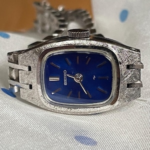 May include: A silver wristwatch with a blue face and a silver metal band. The watch face has the word "SEIKO" printed on it.