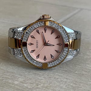 Puede incluir: Un reloj de la marca Seksy con esfera rosa y correa dorada y plateada. La esfera del reloj está rodeada por un anillo de pequeños cristales transparentes. La correa tiene eslabones dorados y plateados. Las manecillas del reloj son negras.