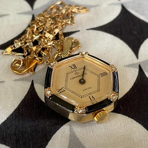 Könnte beinhalten: Eine goldfarbene Anhängeruhr mit sechseckigem Zifferblatt und schwarzen Akzenten. Das Zifferblatt ist cremefarben mit schwarzen römischen Ziffern und dem Aufdruck "Continental". Eine goldene Kette und ein Verschluss sind angebracht.