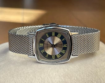 May include: A silver watch with a square face and a mesh metal band. The watch face has black Roman numeral hour markers and a blue second hand. The watch face also has the word "Sekonda" printed on it.