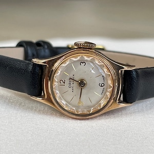 Peut inclure: Une montre-bracelet dorée avec un cadran blanc et un bracelet en cuir noir. Le cadran de la montre comporte des chiffres romains noirs et les mots "ORIS ANTI-SHOCK" imprimés dessus. La montre porte également les mots "SWISS MADE" imprimés sur le cadran.