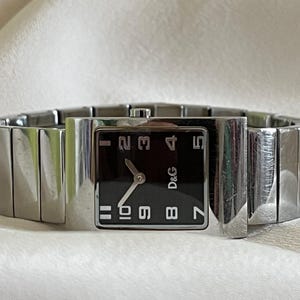 Op de afbeelding: Zilverkleurig polshorloge met een rechthoekige zwarte wijzerplaat met witte cijfers en het D&G-logo. Het horloge heeft een bijpassende zilveren band met een gesegmenteerd ontwerp. Het horloge wordt getoond op een witte ondergrond.