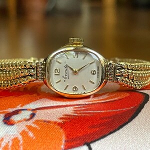Könnte beinhalten: Eine goldfarbene Vintage-Armbanduhr mit cremefarbenem Zifferblatt. Die Uhr trägt den Markennamen "Everite" und die Worte "Swiss Made". Das Armband ist goldfarben mit einem gewebten Design.