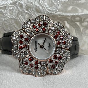 May include: An Ingersoll watch with a flower-shaped face and a black leather strap. The watch features a flower design with clear and red gemstones. The watch face is white with black hands.