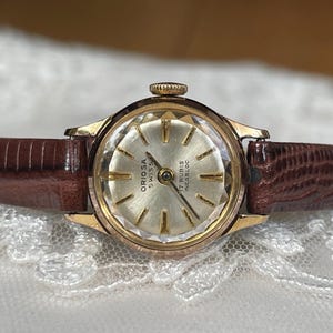 May include: A vintage Oriorsa Swiss watch with a gold-tone case and a textured, light-coloured face. The watch features a brown leather strap. The watch face has the text "ORIOSA SWISS 17 RUBIS INCABLOC".