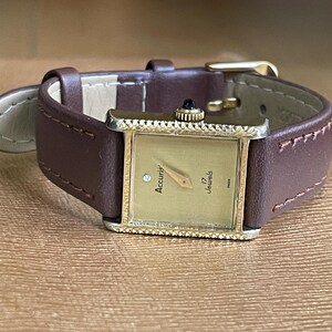 K&ouml;nnte beinhalten: Eine goldfarbene Accurir Armbanduhr mit quadratischem Zifferblatt und braunem Lederarmband. Das Zifferblatt der Uhr hat ein goldfarbenes Zifferblatt mit schwarzen Zeigern und einer rautenf&ouml;rmigen Markierung auf der 12-Uhr-Position. Das Zifferblatt der Uhr tr&auml;gt auch den Text "Accurir" und "17 Jewels".