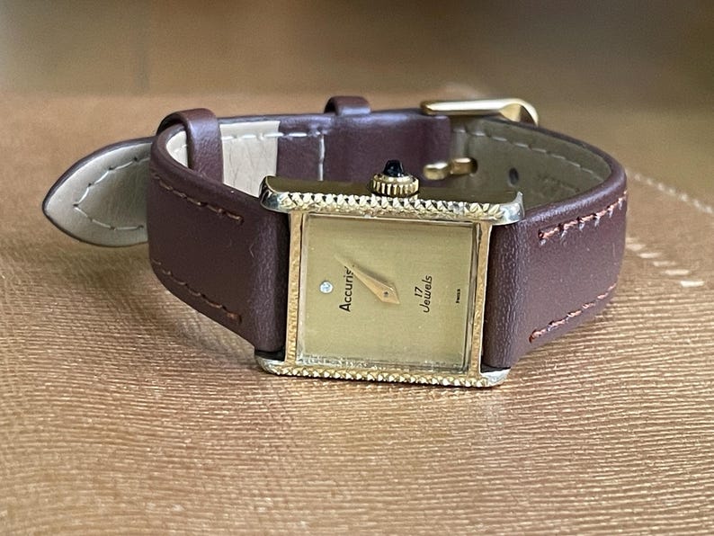 K&ouml;nnte beinhalten: Eine goldfarbene Accurir Armbanduhr mit quadratischem Zifferblatt und braunem Lederarmband. Das Zifferblatt der Uhr hat ein goldfarbenes Zifferblatt mit schwarzen Zeigern und einer rautenf&ouml;rmigen Markierung auf der 12-Uhr-Position. Das Zifferblatt der Uhr tr&auml;gt auch den Text "Accurir" und "17 Jewels".