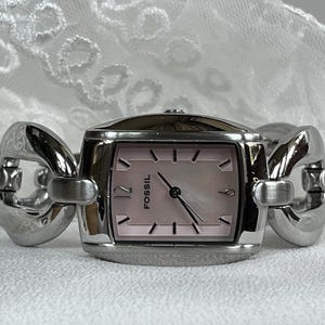 Può includere: Orologio da polso Fossil con quadrante rettangolare rosa e cinturino in metallo color argento. L'orologio ha un quadrante rosa chiaro con lancette e indici neri. Il cinturino presenta un design a maglie.