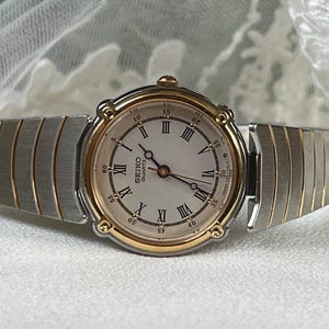 May include: A Seiko Quartz wristwatch with a silver and gold-tone case and strap. The watch face is light-coloured with black Roman numerals and hands. The strap is a flexible, expanding style.