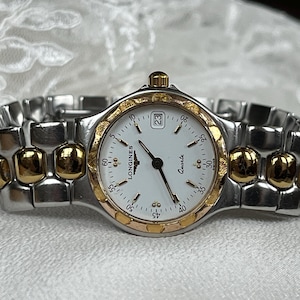 May include: A Longines quartz watch with a silver and gold-tone metal band. The watch face is white with black hands and numerals, and a date display at the 3 o'clock position. The watch has a gold-tone bezel and crown.