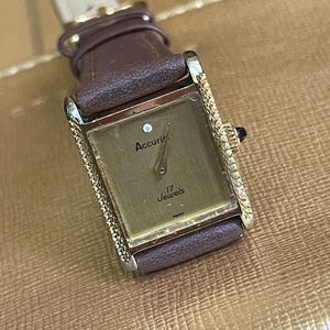 K&ouml;nnte beinhalten: Eine goldfarbene Accuris Armbanduhr mit einem braunen Lederarmband. Das Zifferblatt der Uhr ist rechteckig und hat eine rautenf&ouml;rmige Markierung auf der 12-Uhr-Position. Das Zifferblatt der Uhr zeigt auch "17 Jewels".
