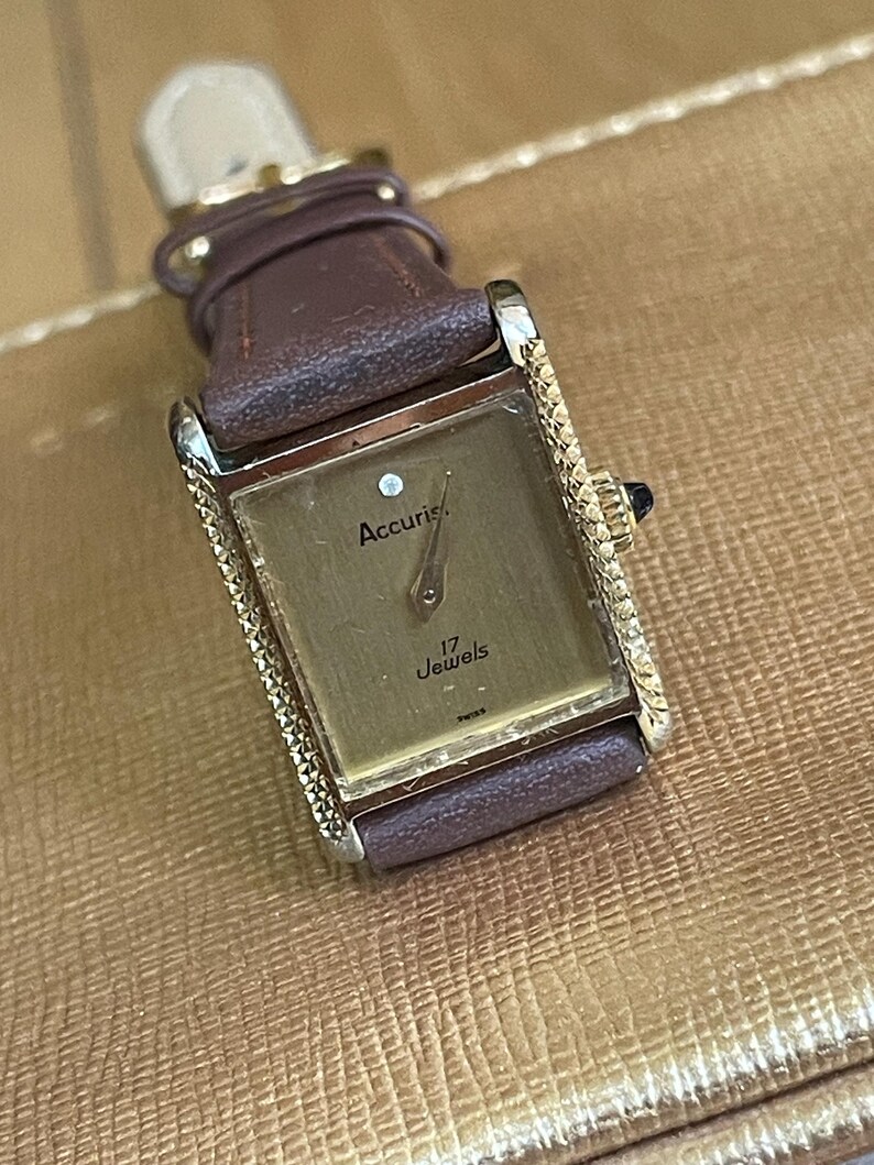 K&ouml;nnte beinhalten: Eine goldfarbene Accuris Armbanduhr mit einem braunen Lederarmband. Das Zifferblatt der Uhr ist rechteckig und hat eine rautenf&ouml;rmige Markierung auf der 12-Uhr-Position. Das Zifferblatt der Uhr zeigt auch "17 Jewels".