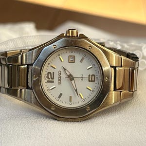 Puede incluir: Un reloj de pulsera Seiko con bisel y correa dorados. La esfera del reloj es blanca con manecillas e indicadores dorados. El reloj muestra los números 12, 6 y 11. La correa está hecha de eslabones rectangulares.