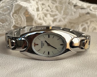 Könnte beinhalten: Silberne und goldene Tissot Armbanduhr mit ovalem Zifferblatt und silbernem Armband. Das weiße Zifferblatt hat schwarze Zeiger und Markierungen. Das Tissot-Logo und "1853" sind sichtbar. Goldakzente an den Seiten.