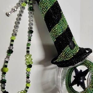 May include: A black and green glass water pipe with a beaded chain attached. The chain has a silver charm with the word "ULTRA" and a silver charm with the word "CHANGE" on it. The pipe has a black and green design with rhinestones.