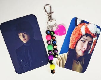 May include: A beaded keychain featuring black letter beads spelling WOODZ, interspersed with colorful star-patterned beads, a bright pink heart charm, and a silver lobster clasp, flanked by two rectangular photo cards of a person in different outfits.