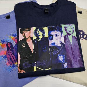 Peut inclure: Trois t-shirts de couleurs bleu et beige. L'un est bleu clair avec un motif éclaboussé coloré et des images de personnes. Le second est bleu marine avec une photo de groupe. Le troisième est beige avec le mot "Rock" en violet.