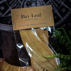 May include: A package of dried bay leaves, labeled "Bay Leaf Apothecary Herbs," with the text "Use for writing intentions or manifestations." The leaves are in a clear plastic bag, resting on a wooden slice, with a dark background.
