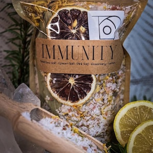 Puede incluir: Una bolsa transparente de sales de baño etiquetada como "IMMUNITY" con rodajas de limón secas y hierbas. La bolsa tiene una etiqueta marrón con el texto "IMMUNITY" y las palabras "Dead Sea Salt - Epsom Salt - Pink Salt - Rosemary - Lemon". La parte superior de la bolsa tiene el texto "Lavender Owl Apothecary".