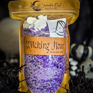 Pode incluir: Uma bolsa transparente contendo sais de banho roxos e pretos, rematada com peças de cera brancas em forma de flor. O rótulo apresenta o texto Lavender Owl Apothecary e Witching Hour Ritual Bath Soak numa fonte preta estilizada.