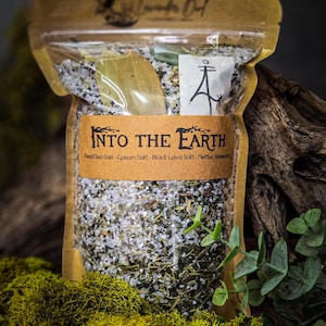 May include: A brown paper bag with a zipper closure filled with a blend of salts and herbs. The bag is labeled "INTO THE EARTH" and lists the ingredients: Dead Sea Salt, Epsom Salt, Black Lava Salt, Nettle, Rosemary.