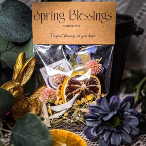 May include: A clear bag filled with dried flowers, orange slices, and herbs, labeled "Spring Blessings Simmer Pot." The bag is attached to a brown tag with the text "Fragrant blessings for your home." A gold bunny figurine and a purple flower are in the foreground.