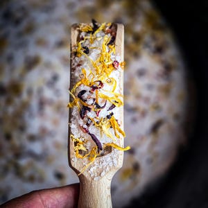May include: A wooden scoop filled with white bath salts, dried orange and red flower petals.