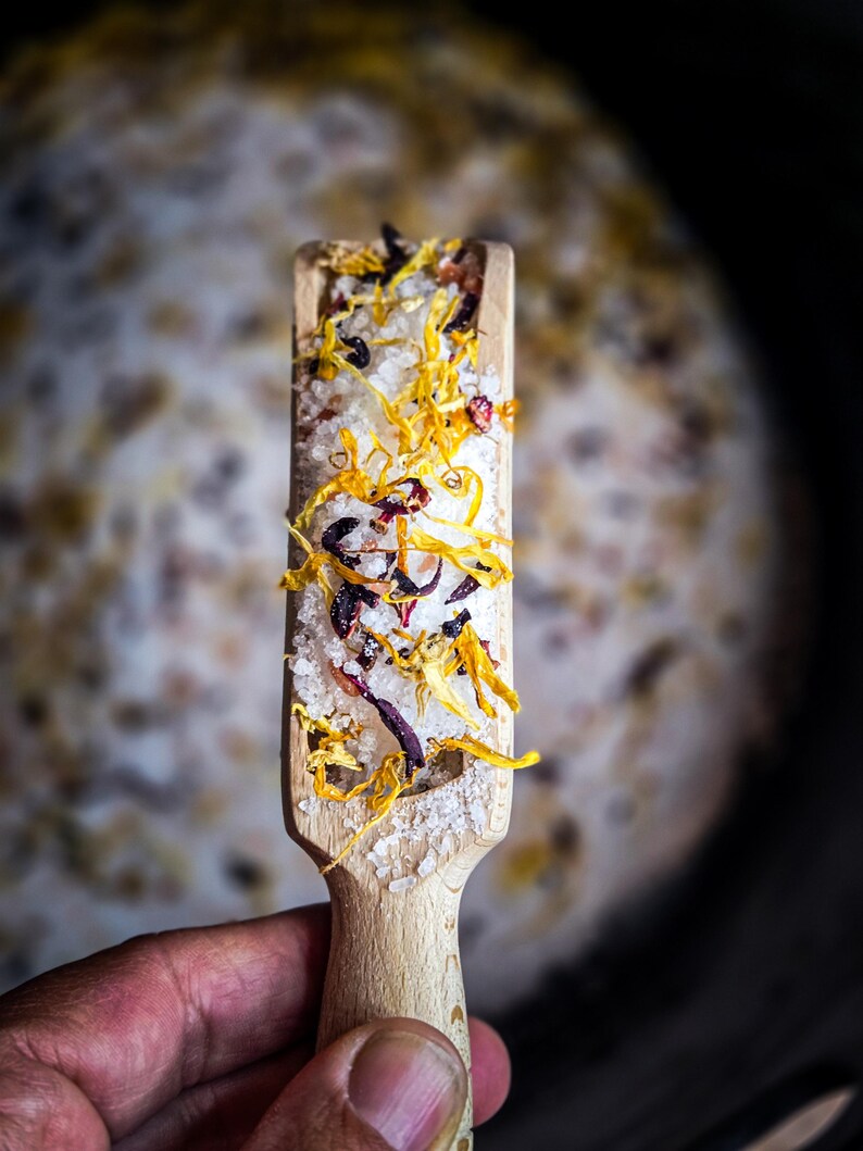 May include: A wooden scoop filled with white bath salts, dried orange and red flower petals.